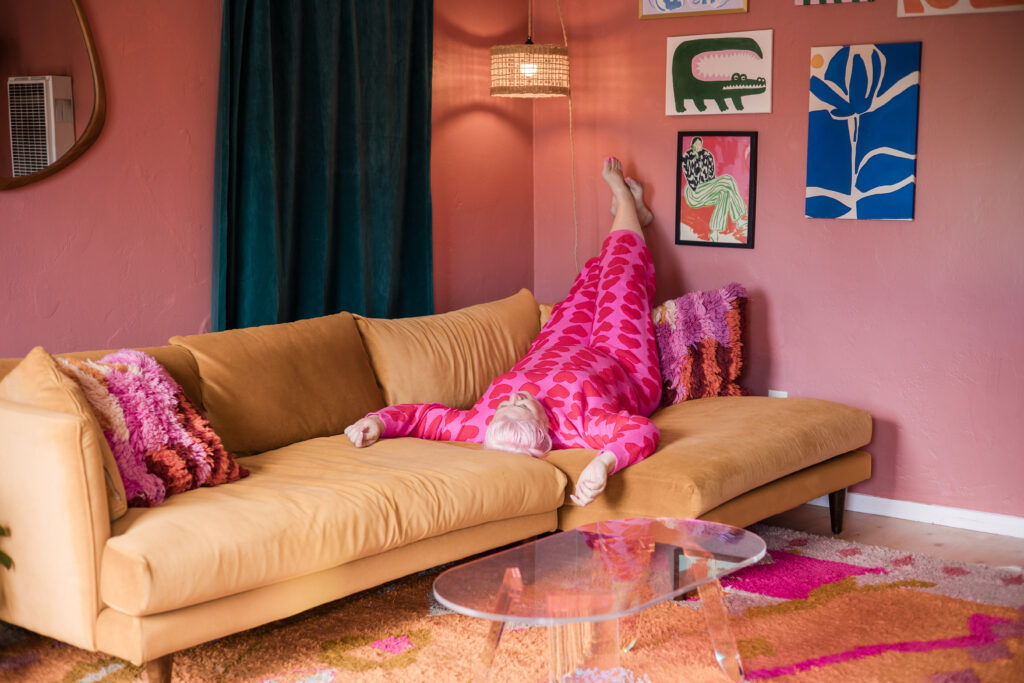 Unison lounges in her pink home, legs up on the wall, taking a well-earned rest after a decluttering session — proof that organizing can feel fabulous.