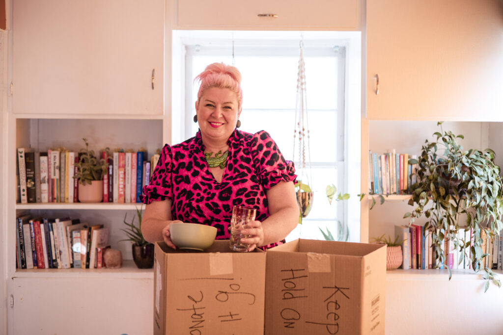 Unison, founder of Go Goddess Organizing, stands in her bright, cozy home, smiling while sorting dishes into boxes labeled “Donate,” “Keep,” and “Let it go” — encouraging women to declutter with confidence, color, and joy.