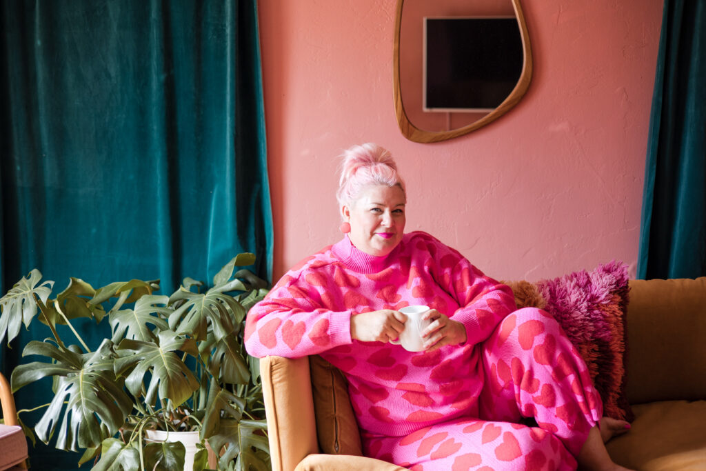 Founder of Go Goddess, Unison, relaxing in a cozy living room with a cup of coffee, wearing a pink heart-patterned sweater and matching pants, surrounded by lush plants and vibrant pillows.