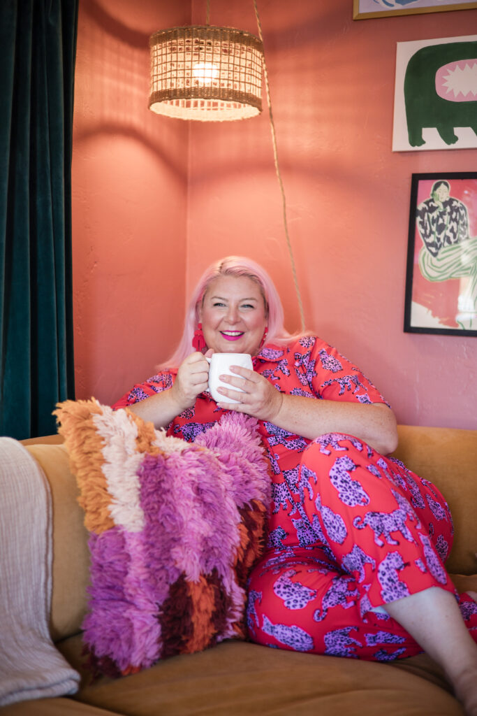 Unison, founder of Go Goddess, smiling and holding a cup of coffee while lounging on a cozy couch in a vibrant pink leopard print outfit, surrounded by colorful pillows and chic décor.