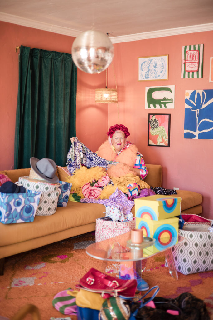 Unison, founder of Go Goddess, surrounded by vibrant fabrics and accessories in a stylish, eclectic living room with colorful décor and a disco ball overhead.