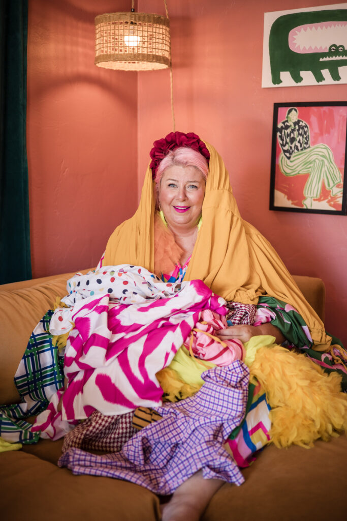 Unison, founder of Unison Creative, sitting on a couch surrounded by colorful patterned fabrics in a vibrant, artistic home studio, creative lifestyle branding and textile design workspace.